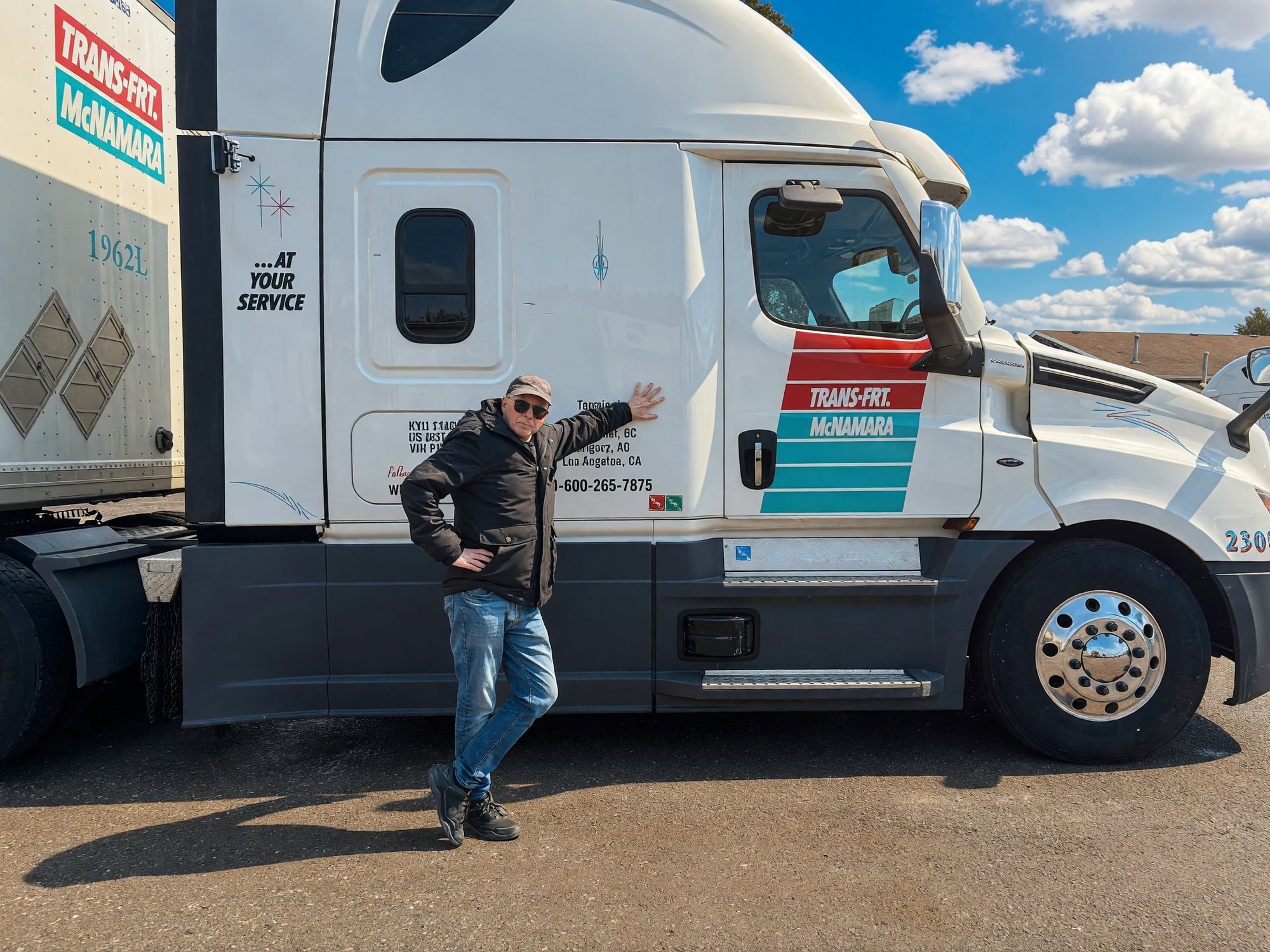 Image shows truck driver for Trans-Frt McNamara posing next to his truck under blue skies and clouds in an open terminal lot. Ayr, Canada. Image shows truck driver for Trans-Frt McNamara posing next to his truck under blue skies and clouds in an open terminal lot. Ayr, Canada.